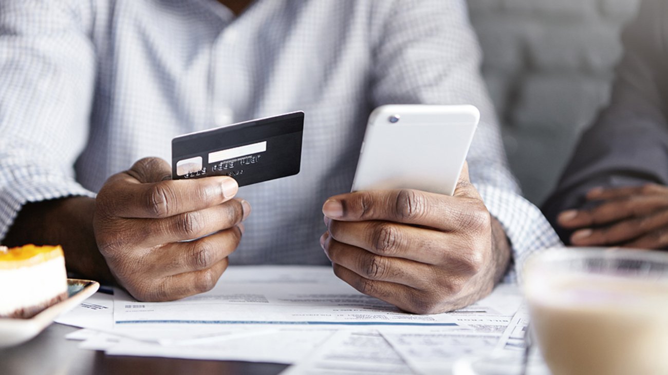 Internet banking, e-commerce and online trading concept. Cropped view of African-American businesman holding mobile phone and credit card while paying bill at cafe. Selective focus on man's hands