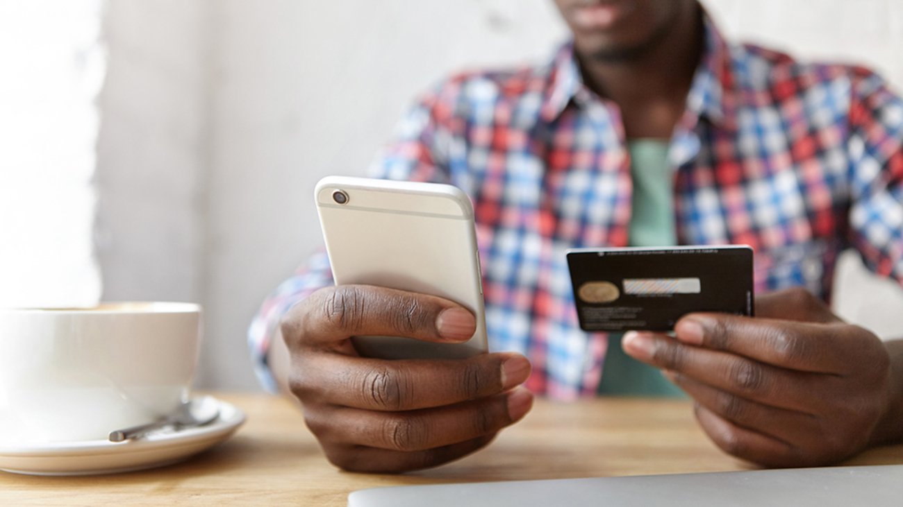 Online payment and shopping. Cropped portrait of African man in plaid shirt holding cell phone in one hand and debit card in other, paying bill at cafe, using mobile banking application during lunch